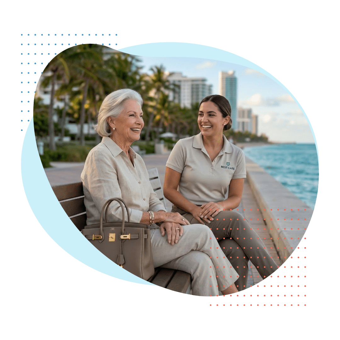 An elegant elderly woman wearing luxury resort wear laughs with a smiling Best Care companion (wearing a discrete polo with a subtle logo), seated on a beachside boardwalk in Bal Harbour, Florida, representing social and refined long-term care.