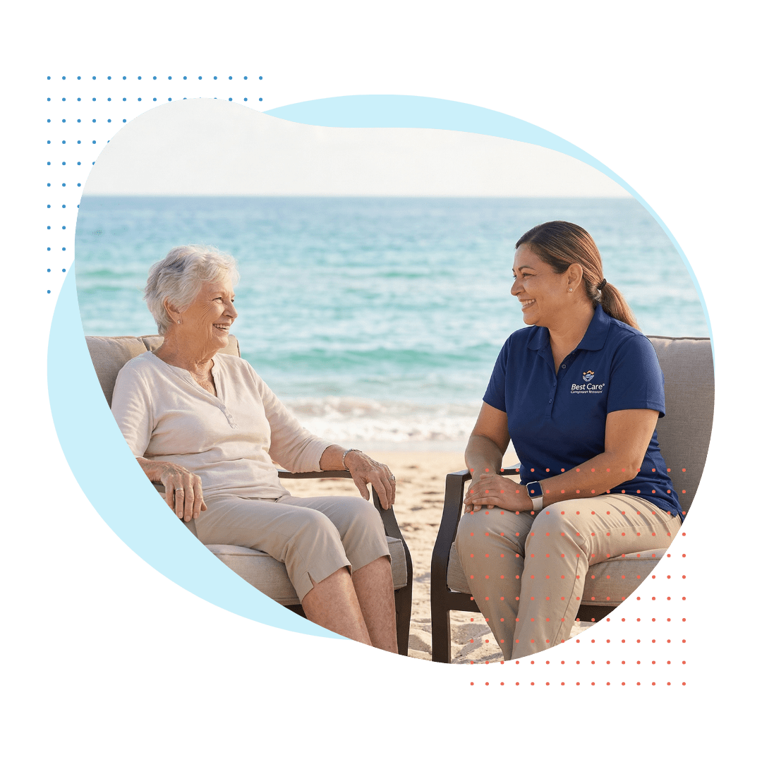 A smiling elderly woman and a friendly female Best Care companion (branded polo shirt and light khakis) are seated in comfortable chairs on the sand, sharing a peaceful moment while looking out at the Atlantic Ocean.